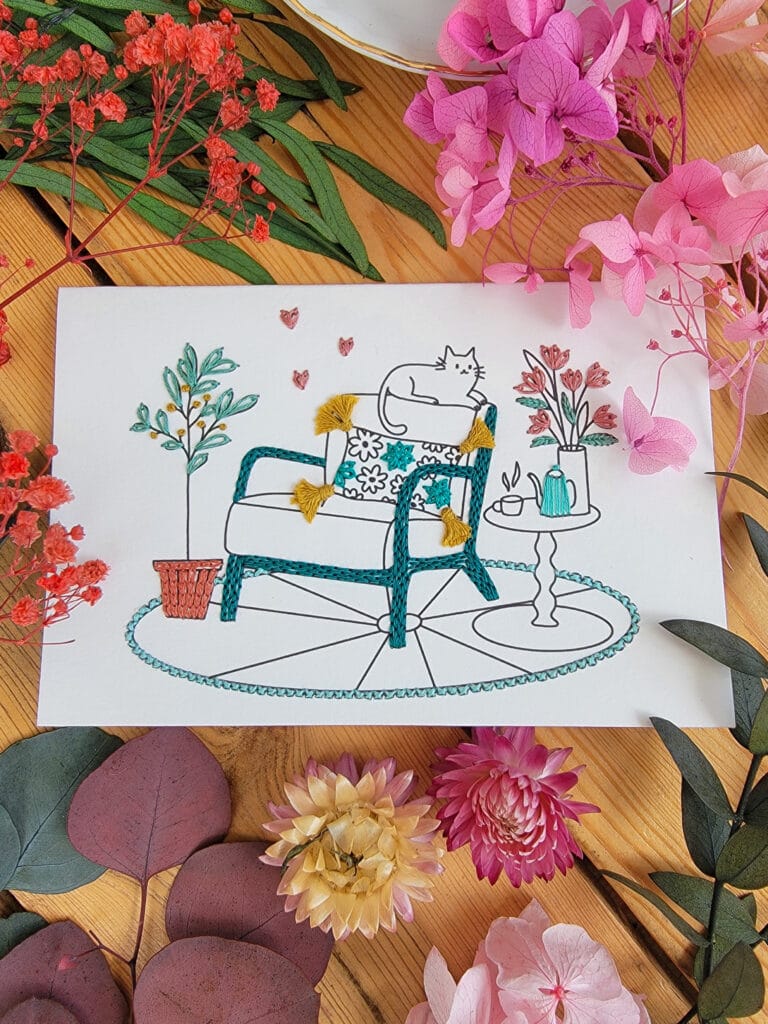 Carte à broder d'un intérieur cocooning : fauteuil relax, chat au repos, et tasse de thé